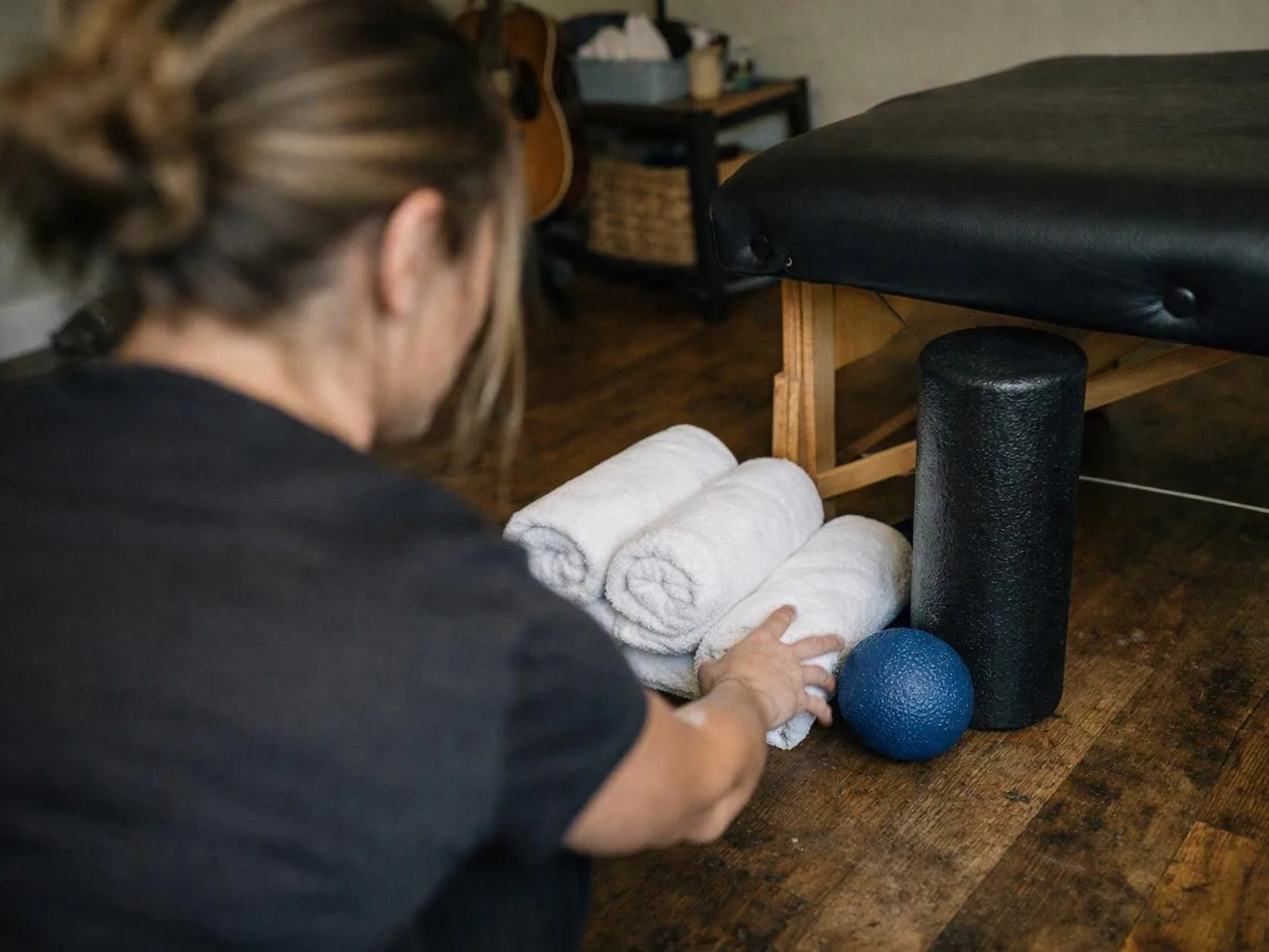 A person reaches toward rolled white towels beside a black massage table and blue massage ball in a room
