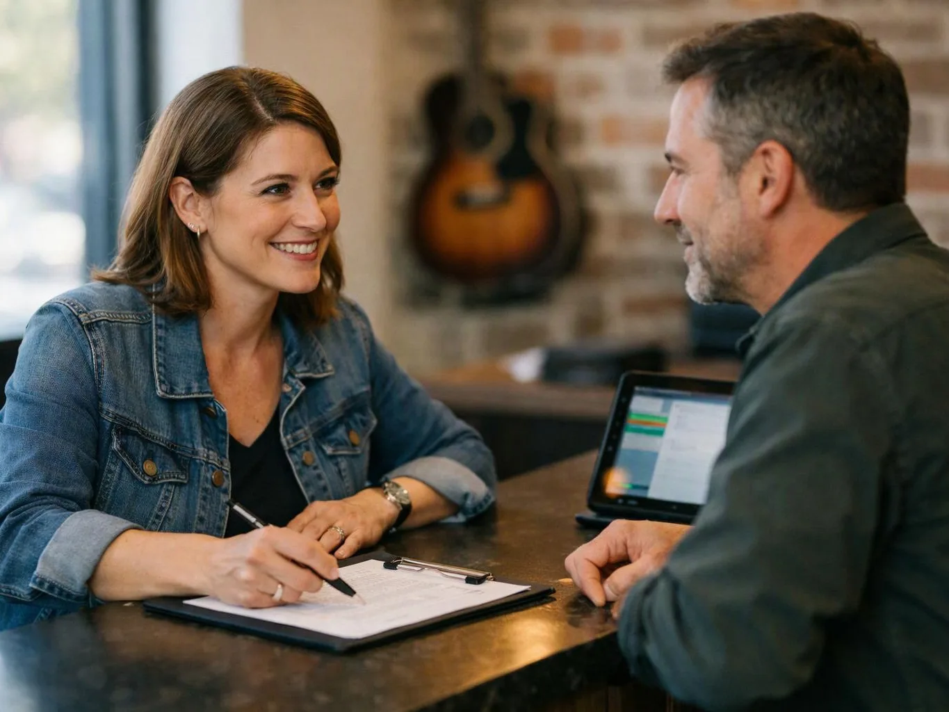 A smiling woman writes on a clipboard while talking with a man across a table in a cozy room with a guitar on the wall